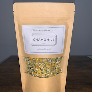 May include: A brown paper bag with a white label that says "Petunia's Herbal Co. Chamomile 1 oz 100% Organic". The bag contains dried chamomile flowers.