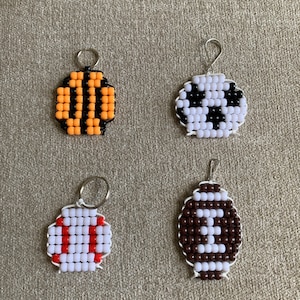 May include: Four beaded keychains with different designs. The first keychain is orange and black with a bee design. The second keychain is white and black with a panda bear design. The third keychain is white and red with a baseball design. The fourth keychain is brown and white with a football design.