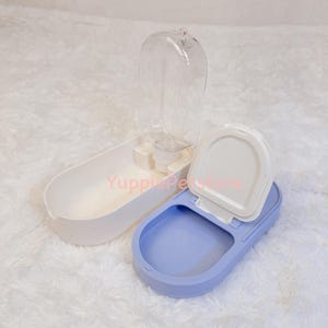 K&ouml;nnte beinhalten: Ein wei&szlig;-blauer Wasserspender und Futternapf f&uuml;r Haustiere. Der Spender hat eine transparente Wasserflasche, ein wei&szlig;es Futtertablett und eine blaue Wasserschale. Die Wasserschale hat einen wei&szlig;en Deckel.