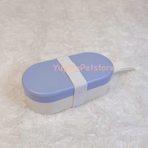 K&ouml;nnte beinhalten: Eine zweifarbige, ovale Lunchbox mit hellblauem Deckel und wei&szlig;em Boden. Ein wei&szlig;er Riemen sichert den Deckel, und ein wei&szlig;er Griff ist angebracht. Die Lunchbox ist zum Transport von Lebensmitteln konzipiert.