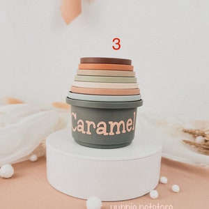 May include: A set of 7 stacking cups in various shades of brown, green, and beige. The cups are made of silicone and have a smooth, rounded finish. The largest cup has the word "Caramel" written on it in pink lettering. The cups are stacked on a white pedestal.