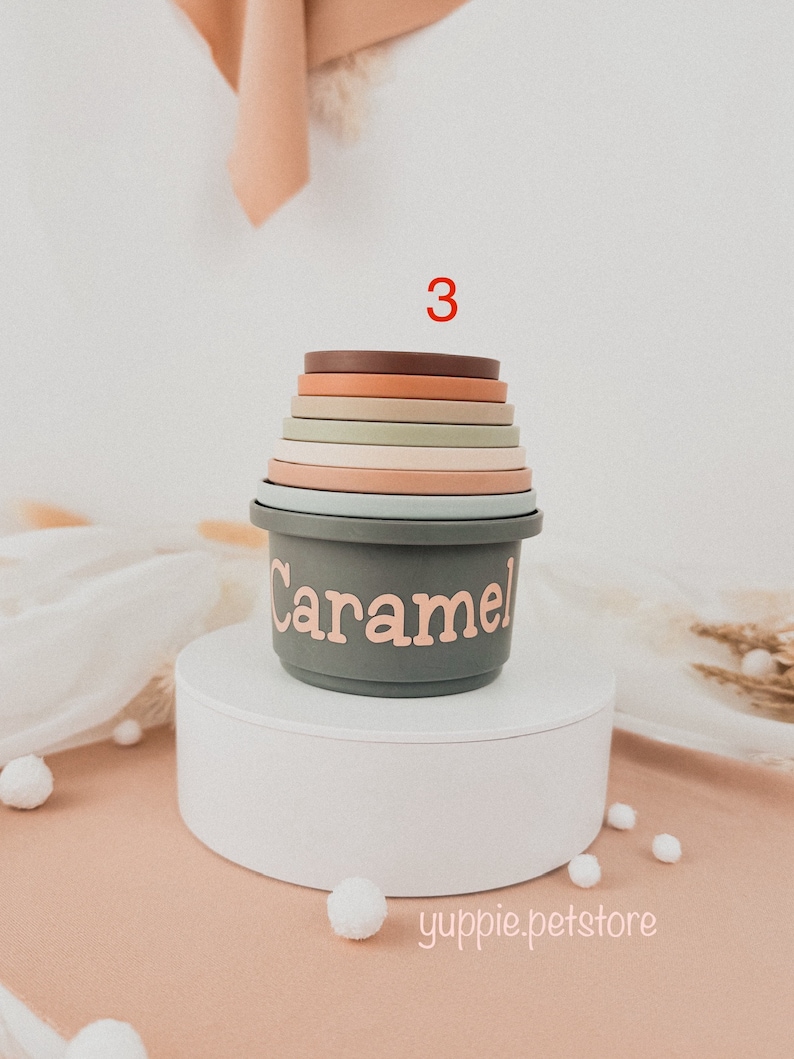 May include: A set of 7 stacking cups in various shades of brown, green, and beige. The cups are made of silicone and have a smooth, rounded finish. The largest cup has the word "Caramel" written on it in pink lettering. The cups are stacked on a white pedestal.