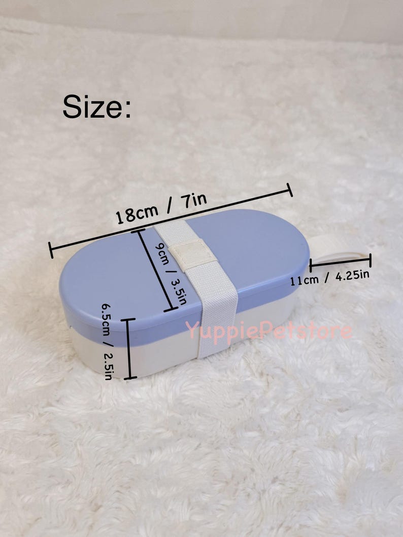 K&ouml;nnte beinhalten: Eine zweifarbige Lunchbox mit hellblauem Deckel und wei&szlig;er Basis. Die Lunchbox ist mit einem wei&szlig;en Riemen gesichert. Abmessungen: 18 cm lang, 9 cm breit und 6,5 cm hoch. Der Text "Size:" ist sichtbar.