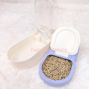 K&ouml;nnte beinhalten: Ein Futter- und Wasserspender-Set f&uuml;r Haustiere. Das Set beinhaltet eine transparente Plastikwasserflasche, einen hellvioletten Futterbeh&auml;lter mit braunen Pellets und eine wei&szlig;e Futterschale. Der Text "Yuppie Petstore" ist sichtbar.