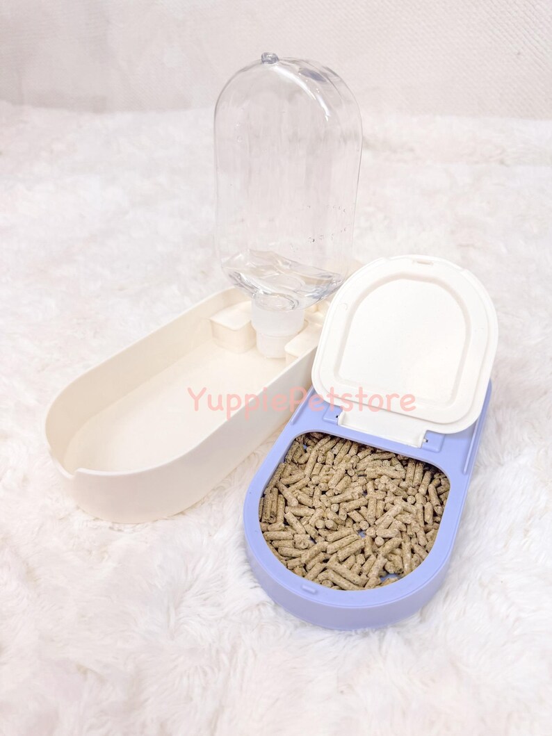 K&ouml;nnte beinhalten: Ein Futter- und Wasserspender-Set f&uuml;r Haustiere. Das Set beinhaltet eine transparente Plastikwasserflasche, einen hellvioletten Futterbeh&auml;lter mit braunen Pellets und eine wei&szlig;e Futterschale. Der Text "Yuppie Petstore" ist sichtbar.