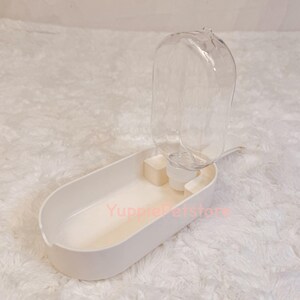 K&ouml;nnte beinhalten: Ein wei&szlig;er Trinkbrunnen f&uuml;r Haustiere mit einer transparenten Plastikwasserflasche. Der Wasserspender hat eine ovale Basis mit einer kleinen Wasserschale und einem transparenten, kuppelf&ouml;rmigen Wasserbeh&auml;lter. Der Text "YuppiePetstore" ist sichtbar.