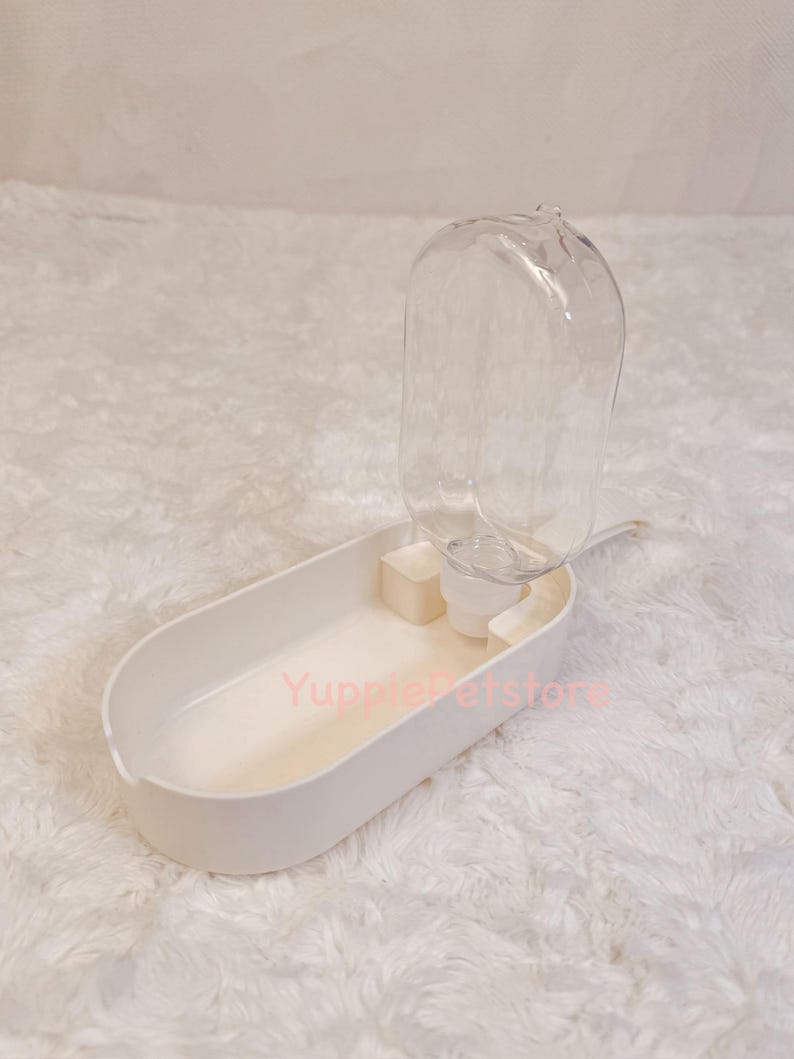 K&ouml;nnte beinhalten: Ein wei&szlig;er Trinkbrunnen f&uuml;r Haustiere mit einer transparenten Plastikwasserflasche. Der Wasserspender hat eine ovale Basis mit einer kleinen Wasserschale und einem transparenten, kuppelf&ouml;rmigen Wasserbeh&auml;lter. Der Text "YuppiePetstore" ist sichtbar.