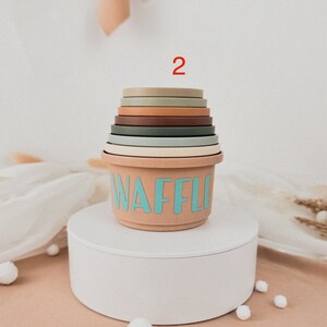 May include: A stackable set of colorful silicone nesting cups with the word "WAFFLE" printed on the largest cup. The cups are stacked on a white pedestal with white pom-poms scattered around the base.