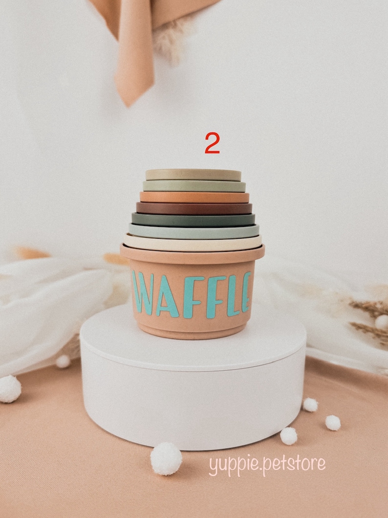 May include: A stackable set of colorful silicone nesting cups with the word "WAFFLE" printed on the largest cup. The cups are stacked on a white pedestal with white pom-poms scattered around the base.