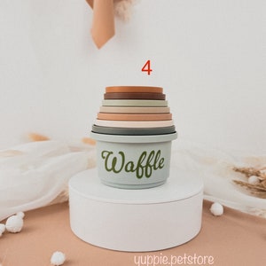 May include: A set of 4 stacking cups in various shades of brown, green, and beige. The cups are made of silicone and have a smooth, rounded finish. The largest cup has the word "Waffle" written on it in green.