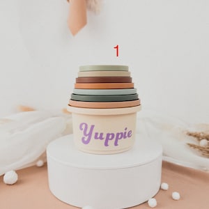 May include: A set of colorful stacking cups with a cream-colored base. The base has the word "Yuppie" written in purple. The cups are stacked in a tower with the number "1" written in red above the tower.