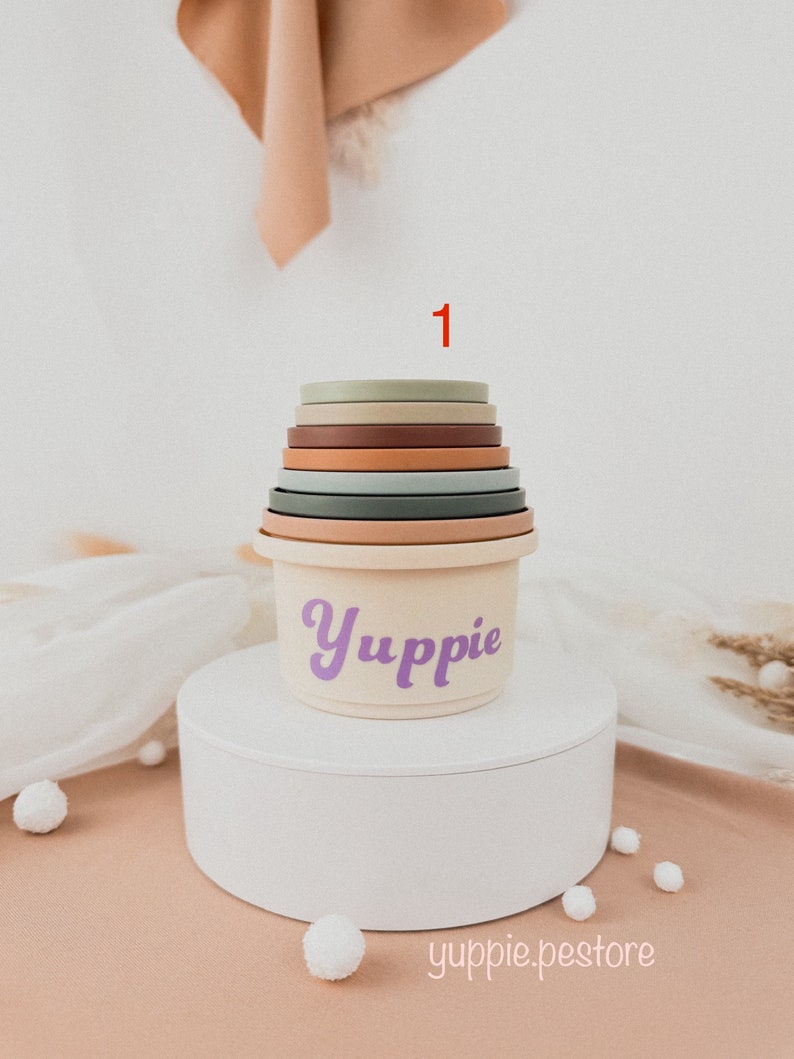 May include: A set of colorful stacking cups with a cream-colored base. The base has the word "Yuppie" written in purple. The cups are stacked in a tower with the number "1" written in red above the tower.