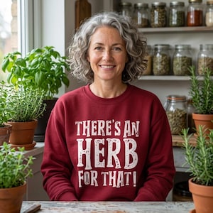 Peut inclure: Un sweat-shirt à manches longues bordeaux avec le texte blanc There's an HERB FOR THAT! imprimé sur le devant. Le vêtement présente une encolure ronde et une coupe décontractée, porté par une personne aux cheveux gris bouclés dans une cuisine.
