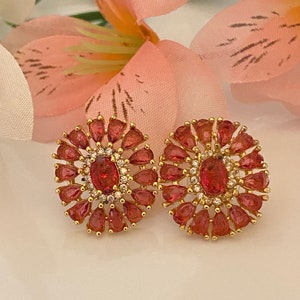 Puede incluir: Un par de pendientes de oro con una piedra preciosa rosa grande de forma ovalada rodeada de piedras preciosas rosas más pequeñas y piedras preciosas redondas transparentes.