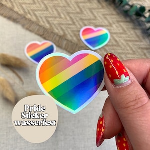 Könnte beinhalten: Drei regenbogenfarbene Herzaufkleber mit irisierendem Effekt und holografischem Finish. Die Aufkleber haben die Form eines Herzens und ein Regenbogenstreifenmuster. Der Text "Pride Sticker wasserfest" ist auf dem Bild zu sehen.