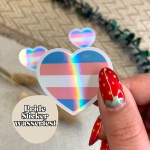 Könnte beinhalten: Ein holografischer Aufkleber in Herzform mit den Farben der Transgender-Pride-Flagge. Der Aufkleber wird von einer Hand mit rotem Nagellack und weißen Punkten gehalten.