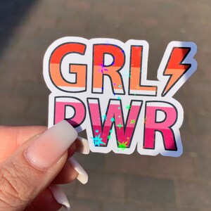 Puede incluir: Una pegatina con las palabras "GRL PWR" en colores arco&iacute;ris con un acabado hologr&aacute;fico. La pegatina tiene un dise&ntilde;o de rayo.