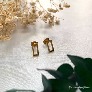 Könnte beinhalten: Ein Paar goldfarbene rechteckige Ohrringe im minimalistischen Design. Die Ohrringe liegen auf einer weißen Oberfläche.