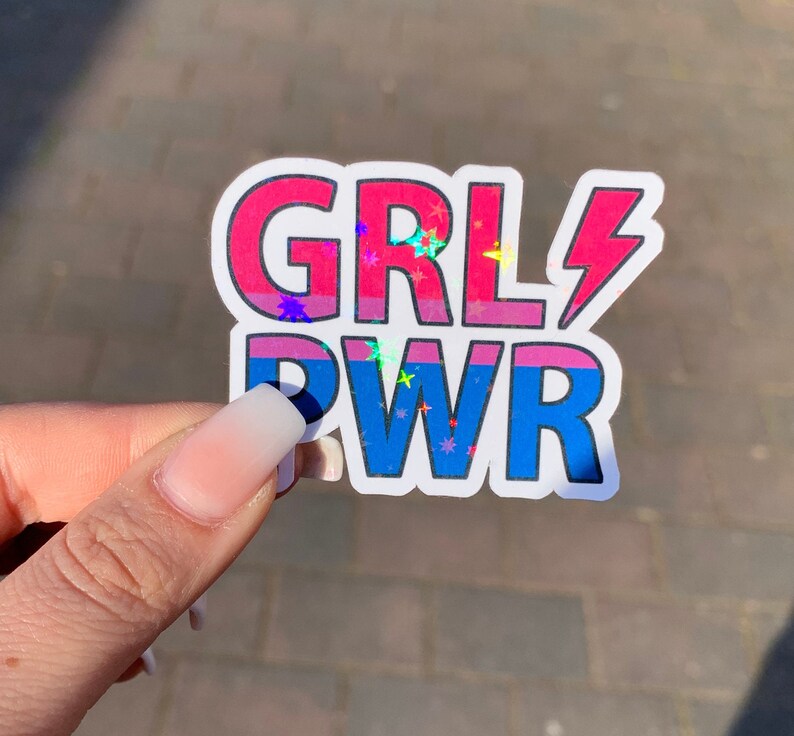 Puede incluir: Una pegatina con el texto "GRL PWR" en rosa y azul con un rayo y estrellas de purpurina.