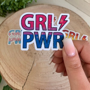 Puede incluir: Una pegatina blanca con el texto "GRL PWR" en rosa, azul y morado con detalles de purpurina. La pegatina tiene un s&iacute;mbolo de rayo junto a la palabra "GRL".