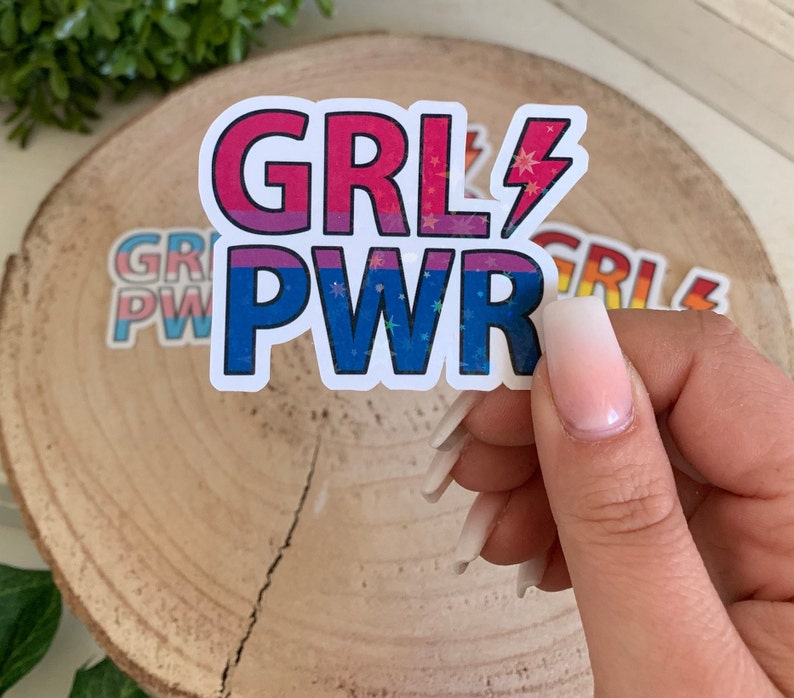 Puede incluir: Una pegatina blanca con el texto "GRL PWR" en rosa, azul y morado con detalles de purpurina. La pegatina tiene un s&iacute;mbolo de rayo junto a la palabra "GRL".