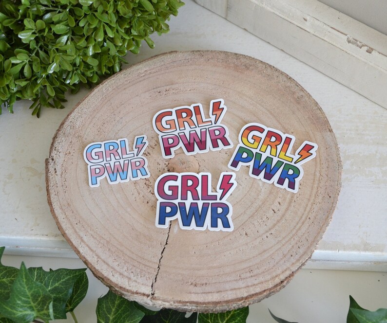 Puede incluir: Cuatro pegatinas de arco&iacute;ris con el texto "GRL PWR" sobre una superficie de madera. Las pegatinas est&aacute;n dispuestas en c&iacute;rculo.