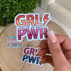 Puede incluir: Tres pegatinas coloridas con el texto "GRL PWR" en rosa, azul y rojo con estrellas blancas. Las pegatinas est&aacute;n sobre una superficie de madera.