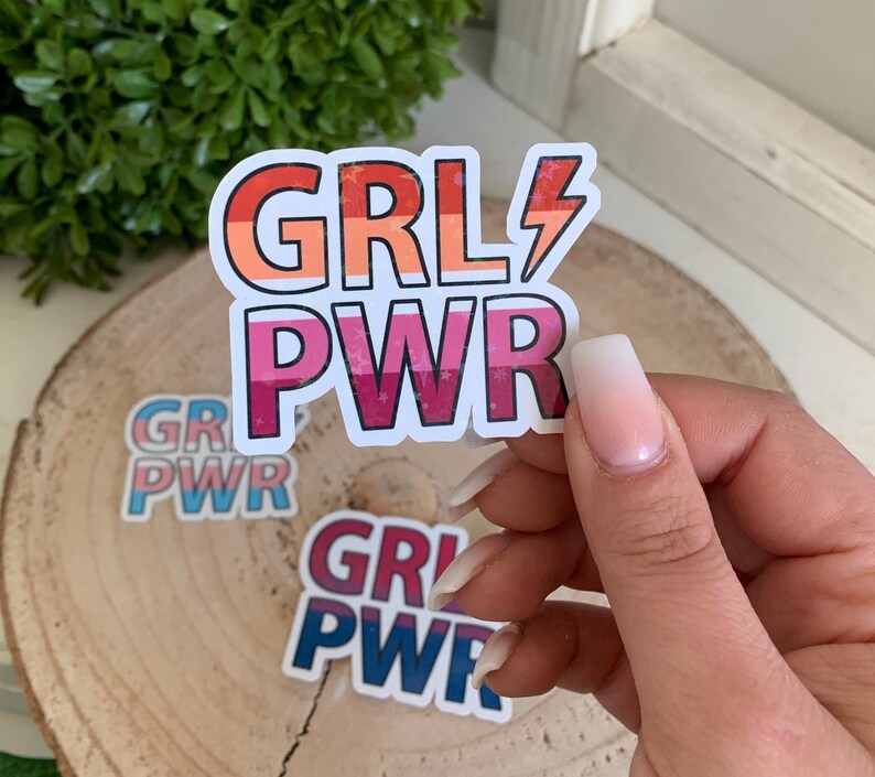 Puede incluir: Tres pegatinas coloridas con el texto "GRL PWR" en rosa, azul y rojo con estrellas blancas. Las pegatinas est&aacute;n sobre una superficie de madera.