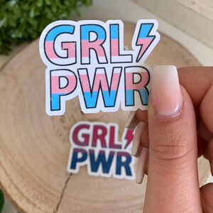 Puede incluir: Dos pegatinas con el texto "GRL PWR" en rayas rosas, azules y blancas con un rayo. La pegatina superior tiene un esquema de colores de la bandera del orgullo transg&eacute;nero y la pegatina inferior tiene un esquema de colores rojo, blanco y azul.