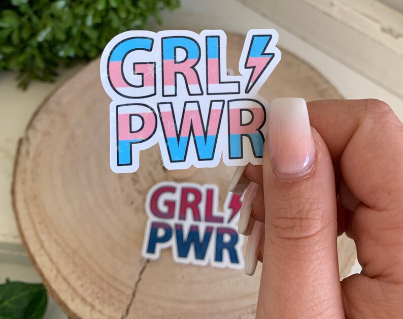 Puede incluir: Dos pegatinas con el texto "GRL PWR" en rayas rosas, azules y blancas con un rayo. La pegatina superior tiene un esquema de colores de la bandera del orgullo transg&eacute;nero y la pegatina inferior tiene un esquema de colores rojo, blanco y azul.