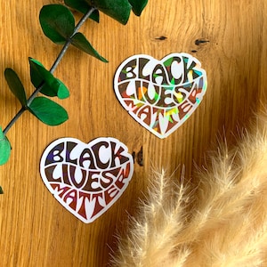 Könnte beinhalten: Zwei regenbogenfarbene Aufkleber in Herzform mit dem Text "Black Lives Matter" in einer stilisierten Schrift.