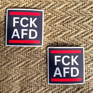 Könnte beinhalten: Zwei schwarze quadratische Aufkleber mit roten Streifen und weißem Text, der "FCK AFD" lautet.
