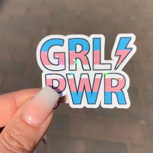 Puede incluir: Una pegatina con el texto "GRL PWR" en letras blancas con un dise&ntilde;o de bandera del orgullo transg&eacute;nero azul y rosa. La pegatina tiene un rayo junto a la palabra "GRL".