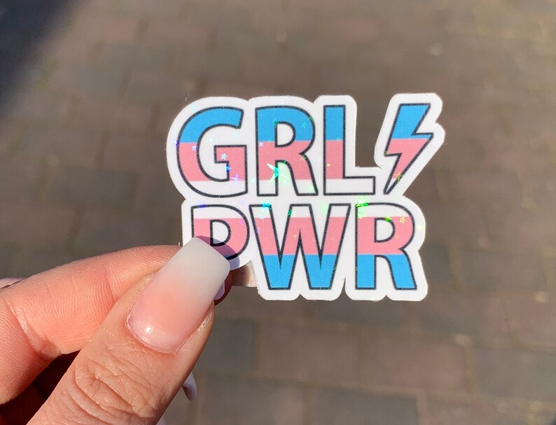 Puede incluir: Una pegatina con el texto "GRL PWR" en letras blancas con un dise&ntilde;o de bandera del orgullo transg&eacute;nero azul y rosa. La pegatina tiene un rayo junto a la palabra "GRL".