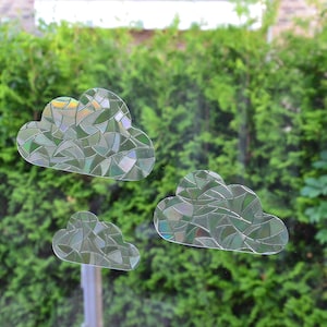 Könnte beinhalten: Drei wolkenförmige Fensteraufkleber mit einem mosaikartigen Design. Die Aufkleber sind transparent mit einer Mischung aus grünen, silbernen und irisierenden Farben. Sie sind an einem Fenster mit einem grünen Heckenhintergrund angebracht.