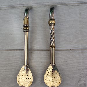 Puede incluir: Dos bombillas de yerba mate. Una tiene un tallo recto plateado con detalles dorados y un extremo dorado perforado en forma de cuchara. La otra tiene un tallo plateado retorcido con detalles dorados.