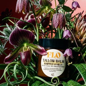 Op de afbeelding: Een pot FLO Tallow Balm Whipped Vanilla, omringd door een weelderig bloemstuk. De bloemen omvatten paarse orchidee&euml;n, gevlekte klokvormige bloemen en donkerrode bloesems. Het etiket van de pot vermeldt de natuurlijke ingredi&euml;nten.