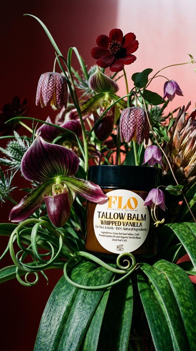 Op de afbeelding: Een pot FLO Tallow Balm Whipped Vanilla, omringd door een weelderig bloemstuk. De bloemen omvatten paarse orchidee&euml;n, gevlekte klokvormige bloemen en donkerrode bloesems. Het etiket van de pot vermeldt de natuurlijke ingredi&euml;nten.