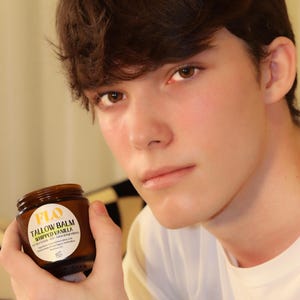 Op de afbeelding: Een bruine glazen pot "FLO Tallow Balm Whipped Vanilla" in de hand van een persoon. De pot heeft een wit etiket met zwarte tekst. De persoon draagt een wit shirt.