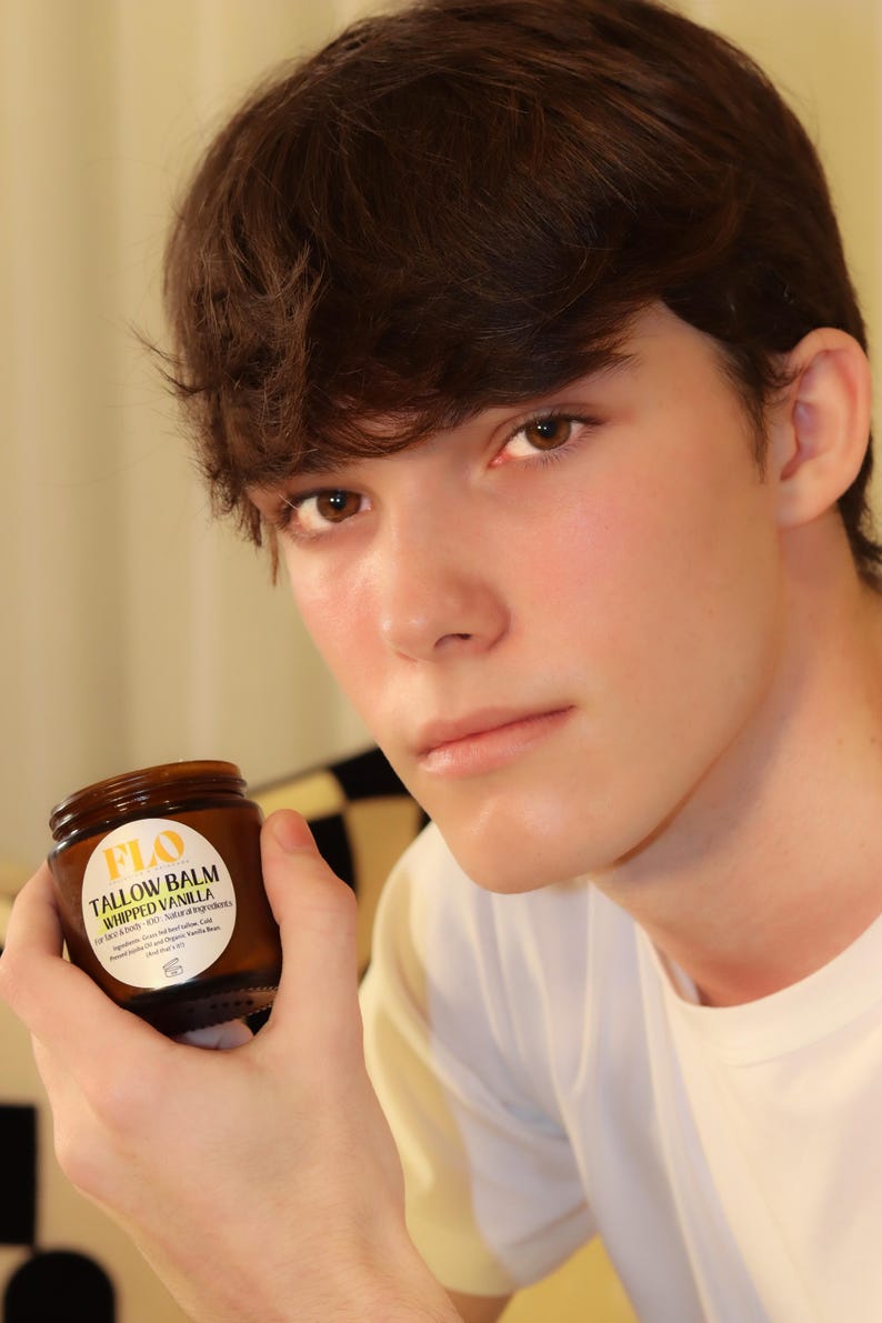 Op de afbeelding: Een bruine glazen pot "FLO Tallow Balm Whipped Vanilla" in de hand van een persoon. De pot heeft een wit etiket met zwarte tekst. De persoon draagt een wit shirt.