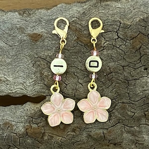 Puede incluir: Dos clips de encanto dorados con encantos de flor de cerezo rosa y una cuenta dorada con una letra negra en cada uno. Los encantos cuelgan de una cadena dorada.