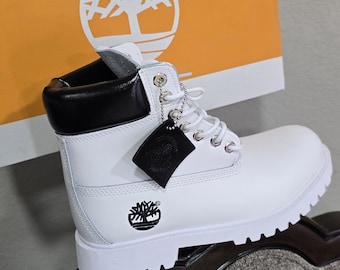 Może przedstawiać: Biały skórzany but z czarnym kołnierzem i białymi sznurowadłami. But posiada logo drzewa Timberland z boku i czarną metkę. Biało-pomarańczowe pudełko z logo Timberland znajduje się w tle.