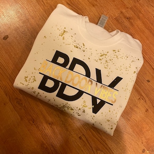 Könnte beinhalten: Weißer Pullover mit einer schwarzen und goldenen Grafik, auf der "Back Door Vibes" mit einem goldenen Sprühmuster steht.