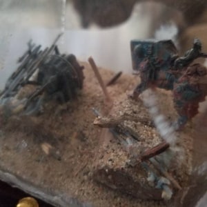May include: A miniature diorama featuring a dark grey mechanical structure and a blue and brown armored figure on a sandy base with scattered wooden sticks, all enclosed within a clear plastic container with a small gold stud visible at the bottom.