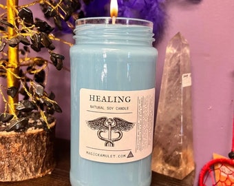 May include: A blue glass candle with a white label that reads "HEALING NATURAL SOY CANDLE" and a medical symbol with a snake wrapped around a staff. The candle is lit and the flame is visible. The text "MAGIC KAMULET.COM" is also on the label.