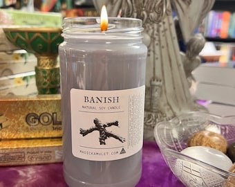 May include: A gray soy candle in a glass jar with a label that reads "Banish Natural Soy Candle" and a stylized image of a stick figure with branches for limbs. The label also includes the website "magickamulet.com".