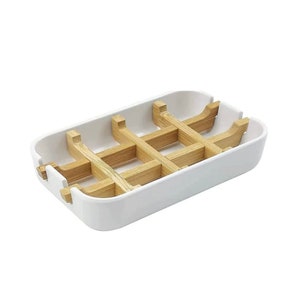 May include: A rectangular white soap dish featuring a removable light brown wooden grid insert. The dish has rounded corners and notched sides to hold the wooden slats in place, creating a tiered structure for drainage and airflow.