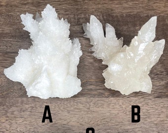 Può includere: Tre gruppi di cristalli bianchi, etichettati A, B e C, ciascuno con una forma e una consistenza uniche. I cristalli hanno un aspetto naturale e ruvido, con bordi affilati e appuntiti. I cristalli sono esposti su una superficie di legno.
