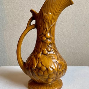 Puede incluir: Jarra de cerámica marrón con asa y boquilla. La jarra presenta un diseño en relieve de uvas y hojas. La jarra está sobre una base redonda y tiene una superficie texturizada.