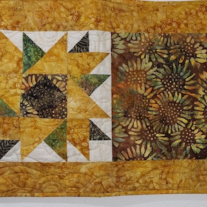 May include: A table runner with a patchwork design featuring a central panel of brown and green floral fabric, flanked by two panels of yellow fabric with a starburst pattern.